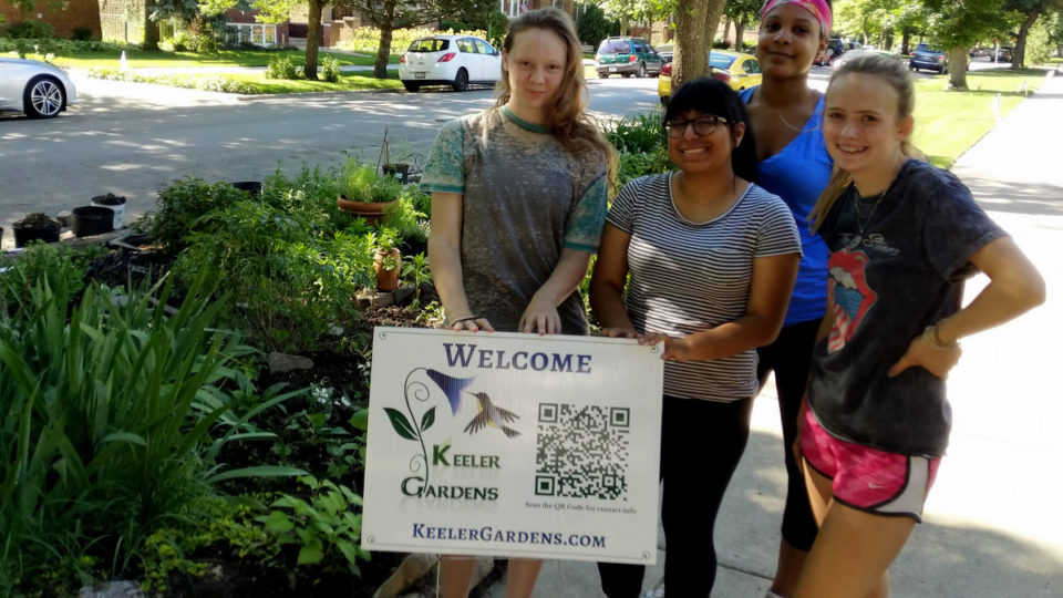 Four high school interns from across Chicago pose for a picture, standing in the right foreground in a group on a sidewalk with a sign that reads “Welcome, Keeler Gardens.” In the background, mostly to the left, is the Keeler Gardens educational pollinator habitat, freshly planted, with a variety of Illinois native pollinator plants.
